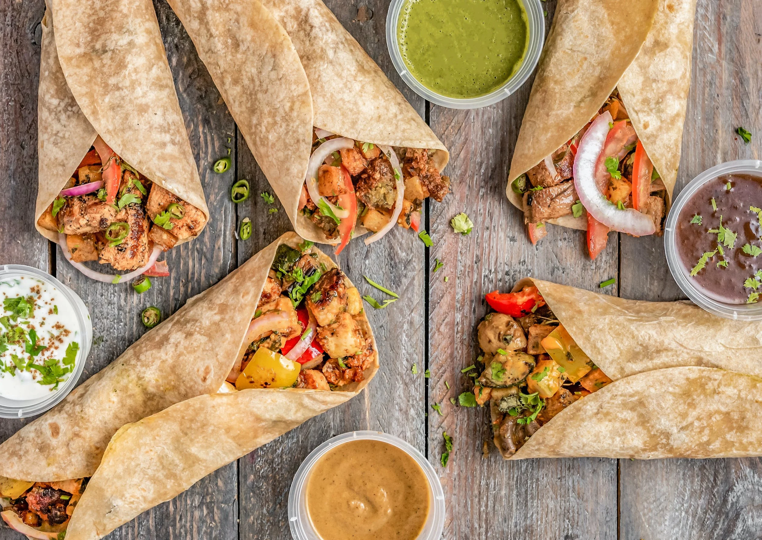 five tacos wrapped in tortillas sit on a wooden table. Green salsa, brown salsa, and orange crema surround the tacos.