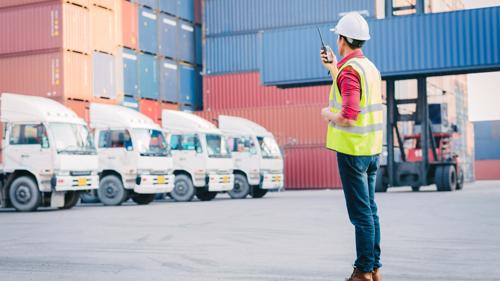 A port dock worker standing at a port talking into. walkie talkie. they are wearing a yellow vest and a white hard hat.