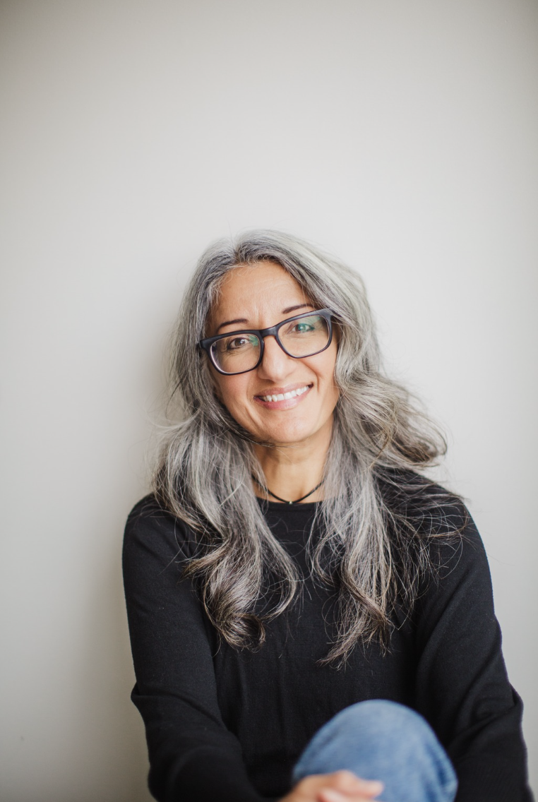 Black and white portrait of a woman with long wavy hair, wearing glasses and a black top, smiling softly against a plain background.