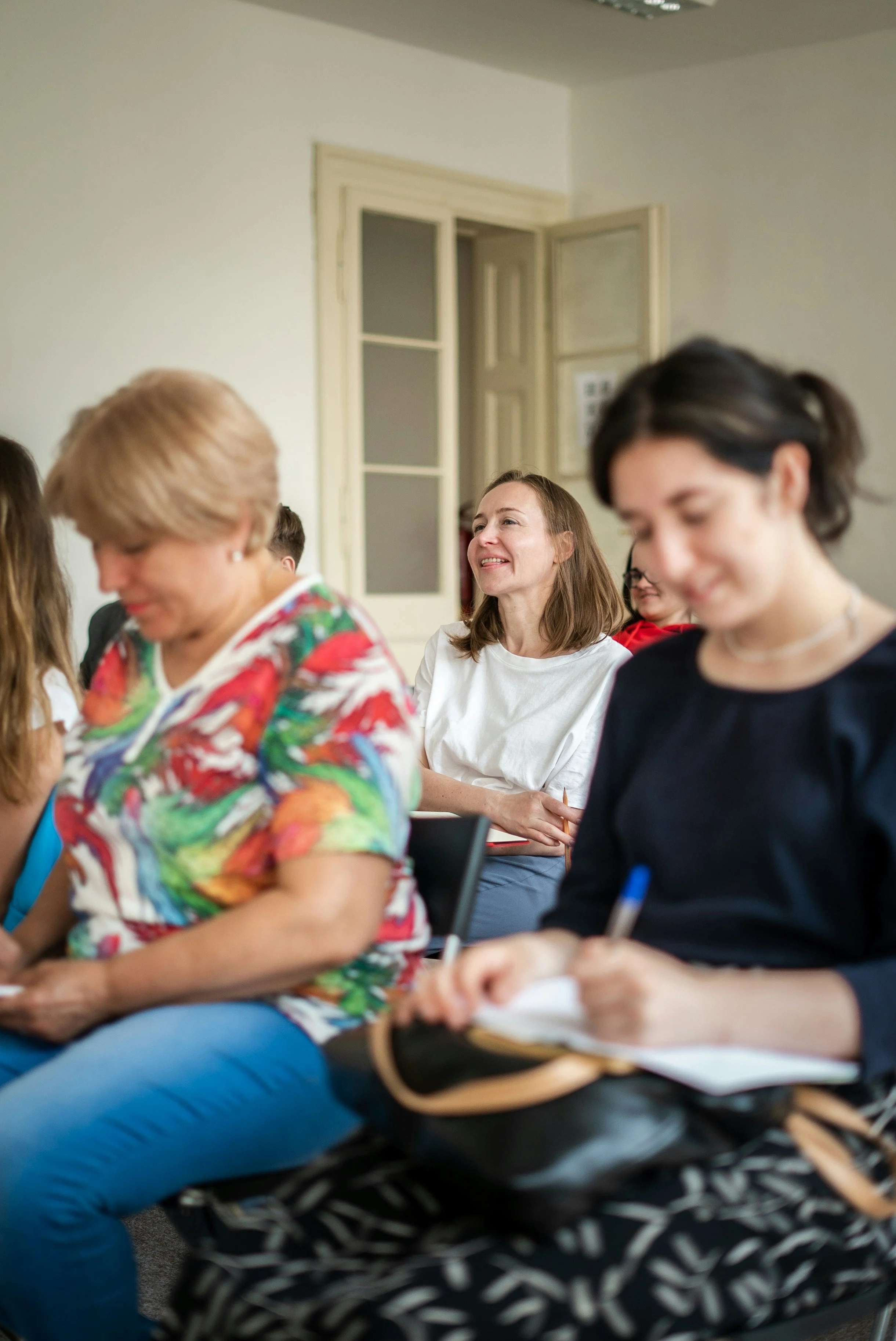 Group of women sitting in a room, attending a meeting or seminar, some taking notes and others smiling.
