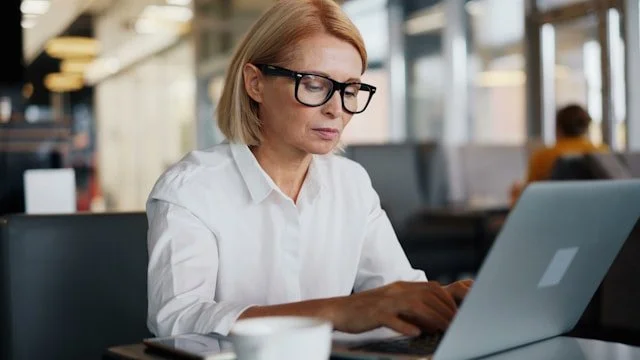 Woman with red hair and glasses working on a laptop in a coffee shop or cafe.
