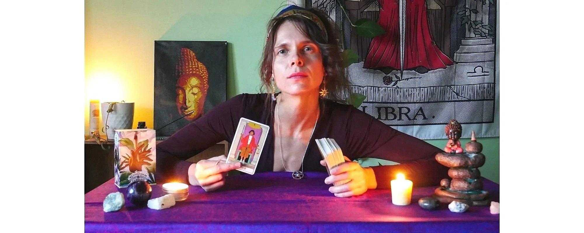 A woman sits at a table with tarot cards, candles, crystals, and a Buddha statue, preparing for a psychic reading.