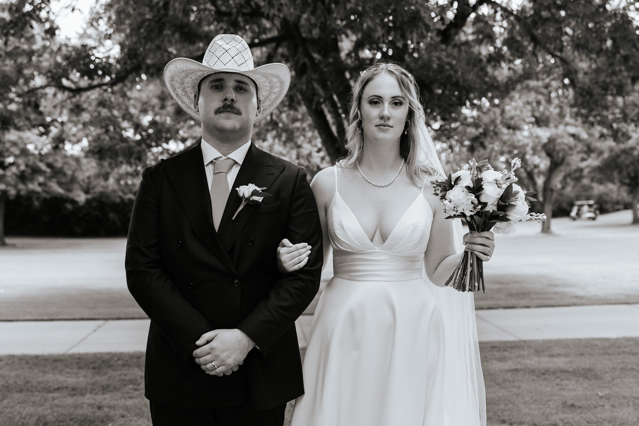 Black-and-white photo of a bride and groom standing together outdoors. The groom is wearing a dark suit with a boutonniere and a large cowboy hat. The bride is in a white wedding dress, holding a bouquet of flowers, and wearing a pearl necklace.