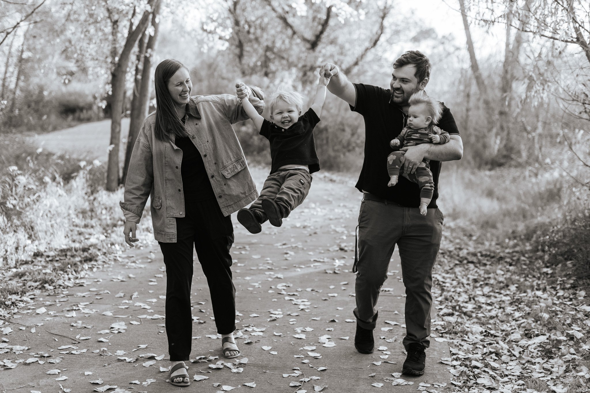 A family of four enjoying a walk outdoors, with a mother, father, and two young children, holding hands and smiling amid trees and fallen leaves.