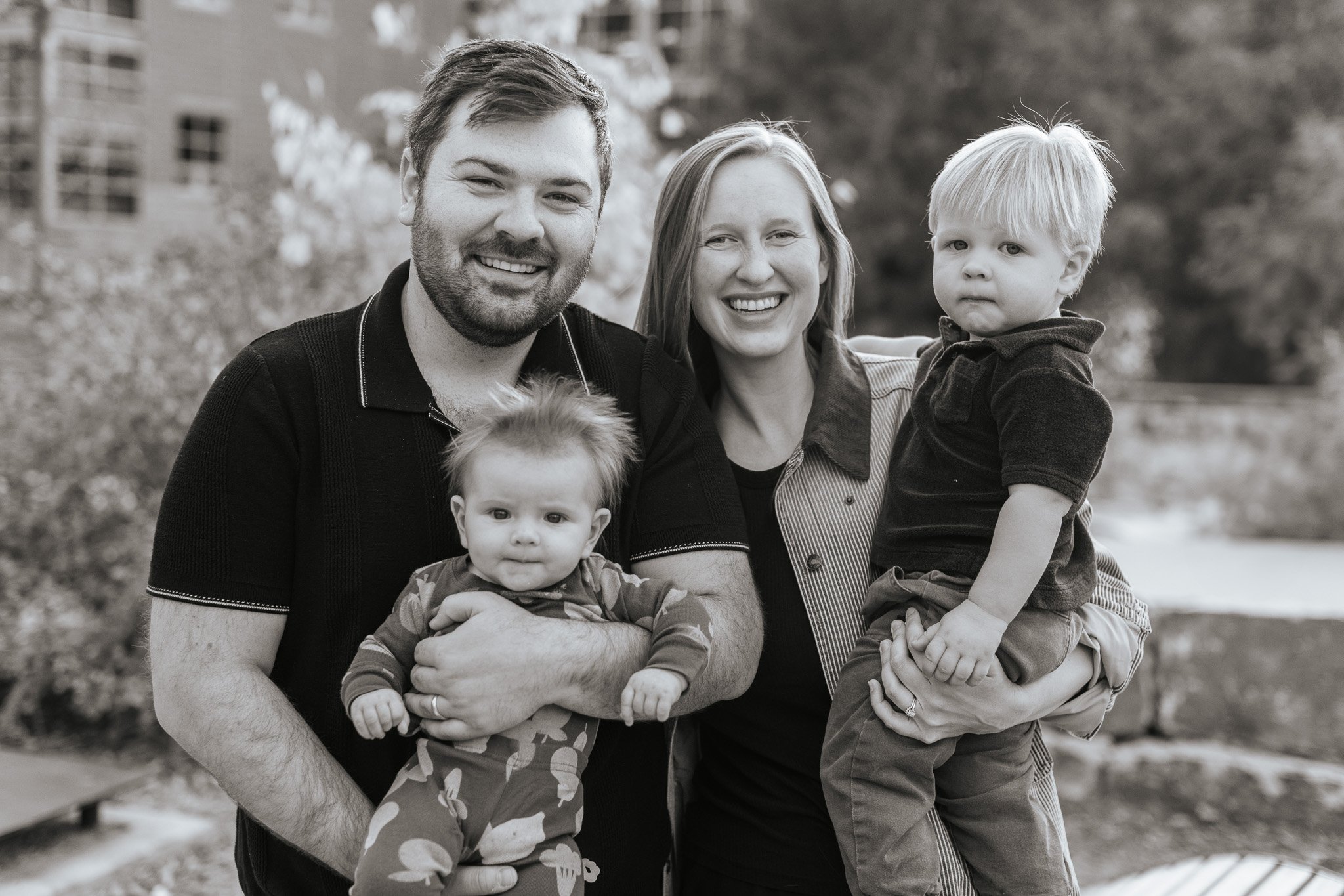 A smiling family of four with two young children outdoors. The father holds a baby girl, and the mother holds a young boy. They are standing close together, with trees and a building in the background.