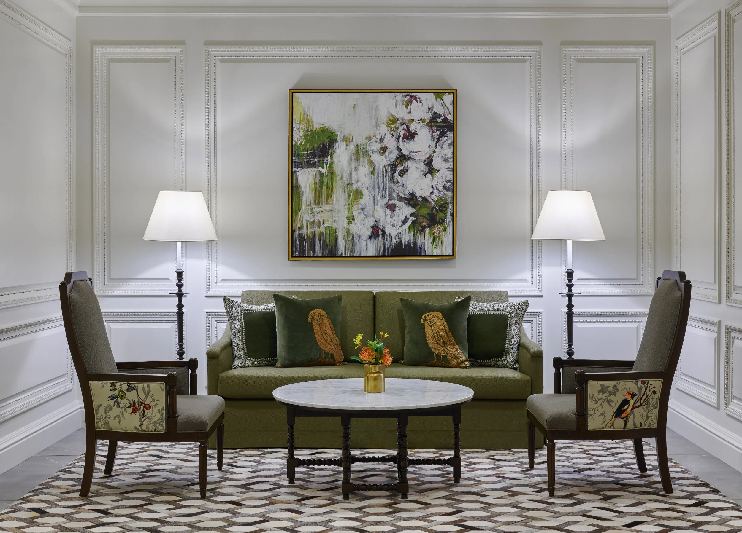 Living room with white paneled walls, green sofa with bird-themed pillows, two patterned chairs with bird embroidery, marble-top coffee table with a small floral arrangement, two floor lamps, and a large abstract painting on the wall.