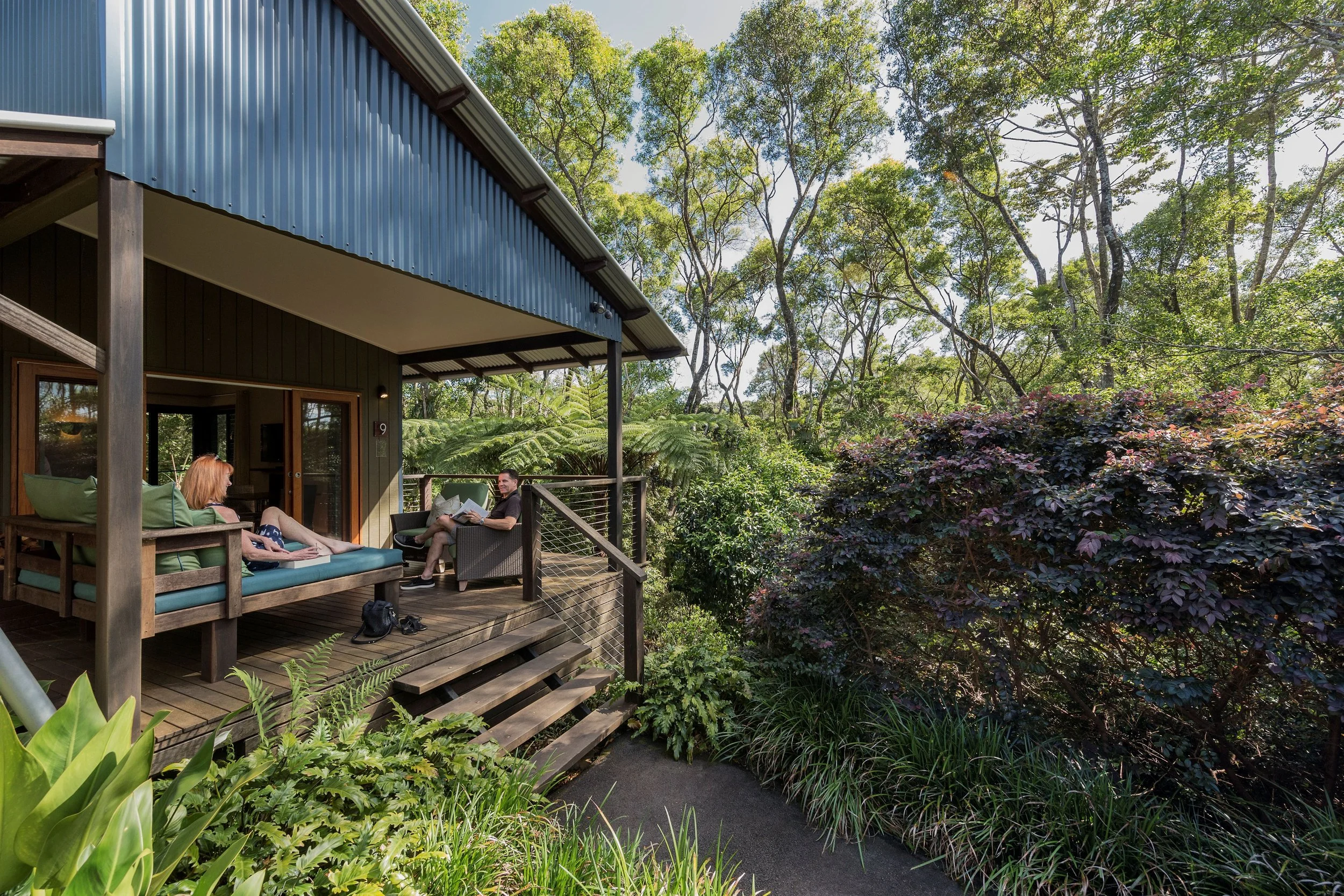 A wooden porch with outdoor seating, two people sitting and talking, surrounded by lush green trees and plants, with a blue and gray metal building nearby.