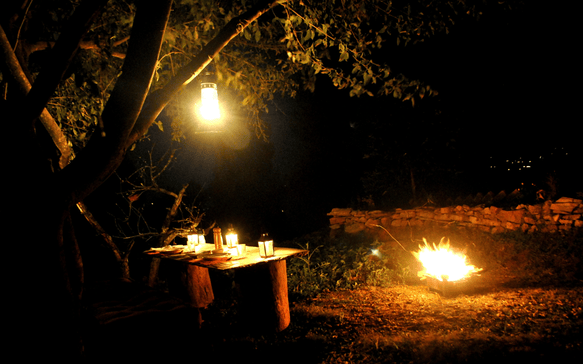 Outdoor dining setup under a tree at night with hanging lantern, candlelit table, and a glowing firepit creating a warm, intimate atmosphere