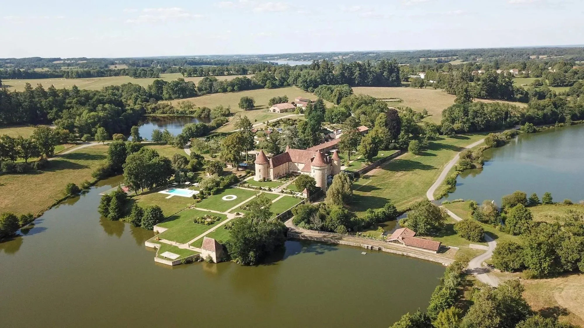 Aerial view of a large estate surrounded by lakes, with a castle-like mansion, manicured lawns, and lush greenery in a rural landscape.