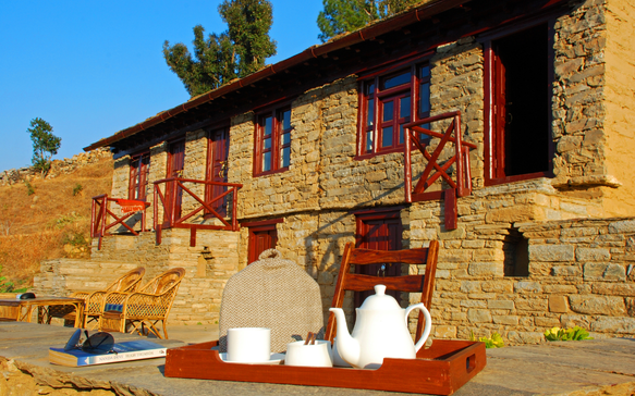 Rustic stone lodge with wooden balconies and outdoor seating, featuring a tea set on a tray in the foreground under clear blue skies