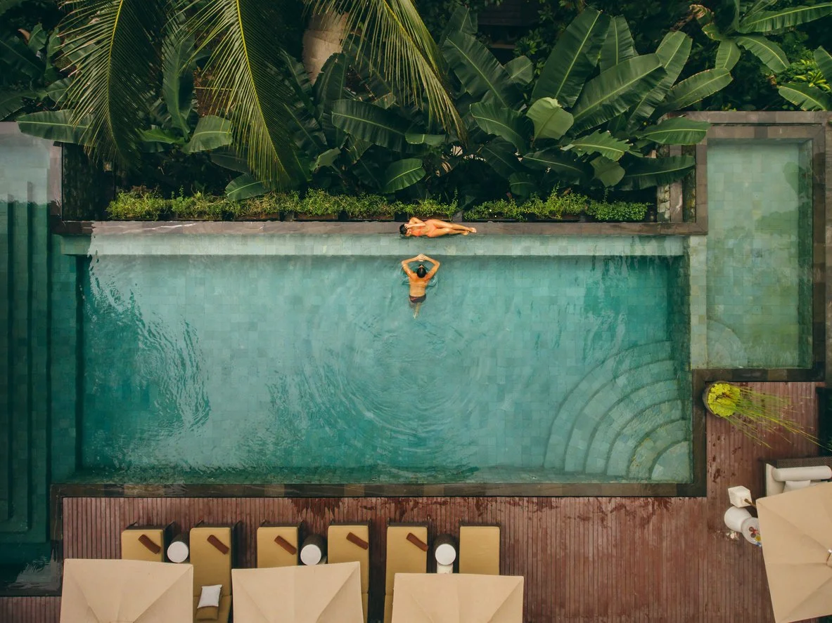 A person swimming in a swimming pool next to a person lying on the poolside with tropical plants in the background.