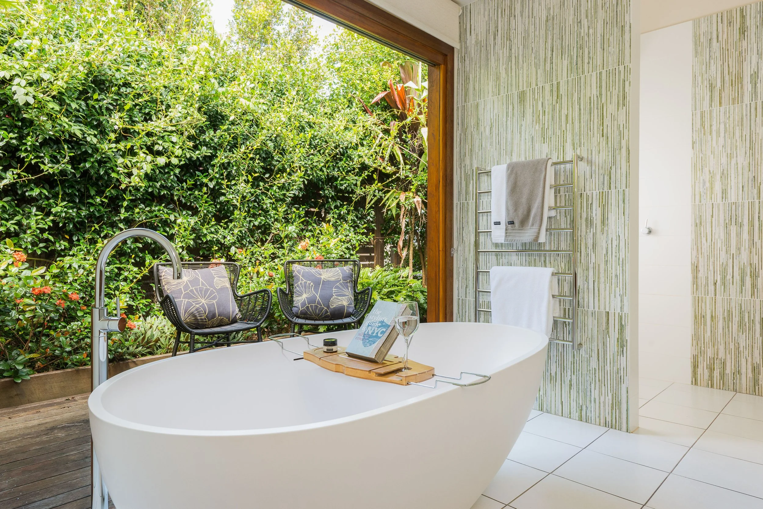 A modern bathroom with a white bathtub, surrounded by a lush green outdoor garden view. The bathroom has patterned green and white tiles on one wall, a towel rack with white towels, and a wooden tray with a book and a glass on the bathtub.
