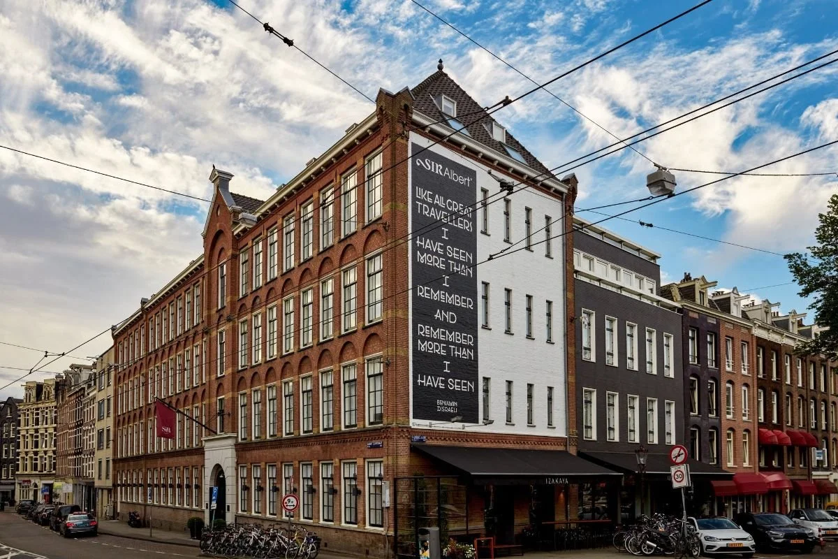 Street view showing a large brick building with a black and white sign, red awnings, bicycles, and parked cars.