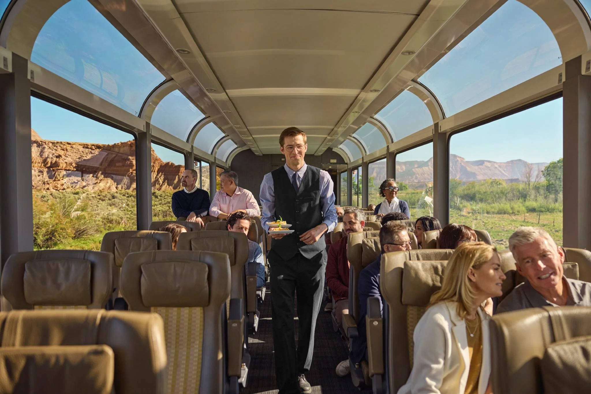 A man in a vest and glasses carrying a plate walks through a bus filled with seated passengers. The bus has large windows showing a desert landscape with cliffs and greenery outside.