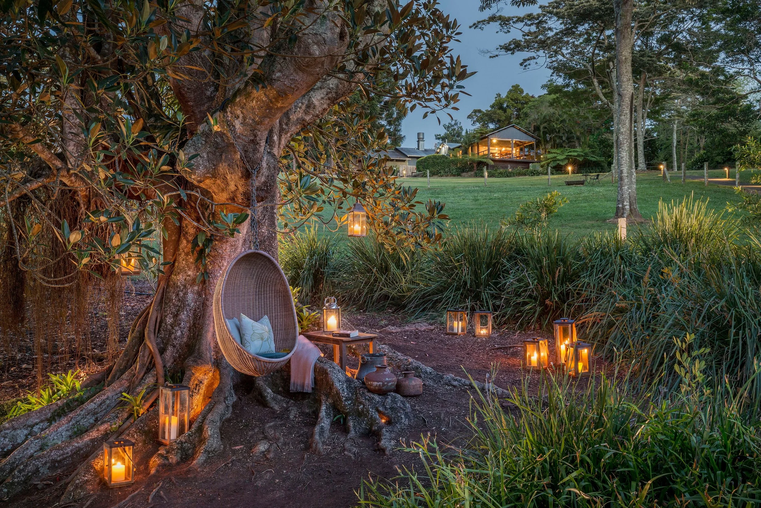 Nighttime outdoor scene with a large tree, illuminated lanterns on the ground, a cozy outdoor chair with cushions, and a house in the background surrounded by trees and greenery.