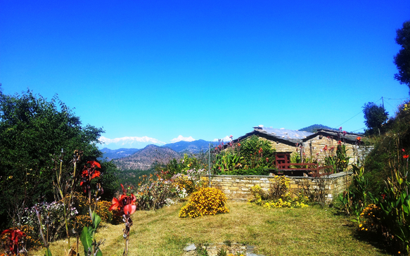 Stone house surrounded by a colourful garden with flowers and greenery, overlooking mountain ranges under a clear blue sky