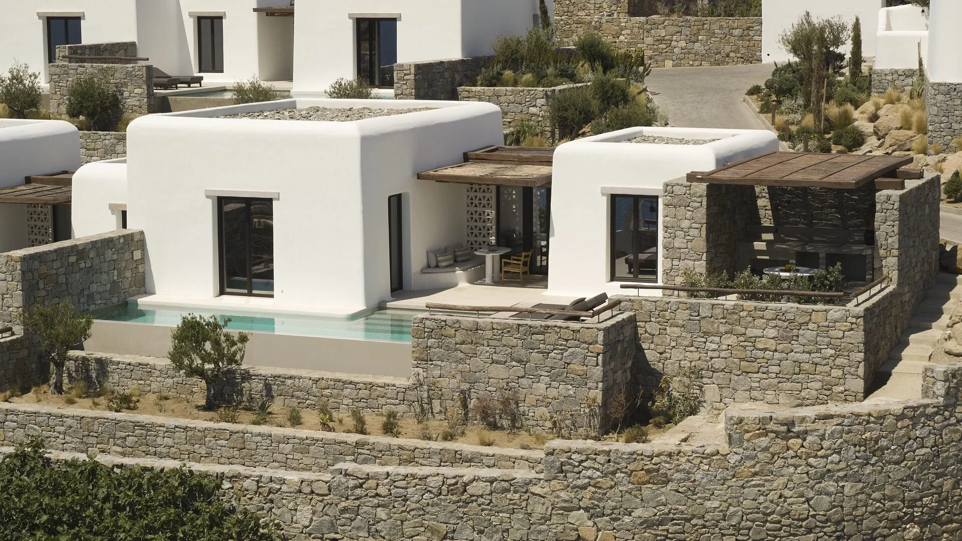 A modern white house with rounded edges, stone walls, and wooden pergolas, situated on a terraced hillside with a small pool and outdoor seating area.