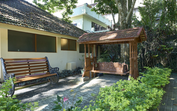 Peaceful Sri Lankan Ayurvedic retreat courtyard with a traditional wooden swing pavilion, shaded garden seating, tiled-roof wellness buildings, and lush tropical greenery.