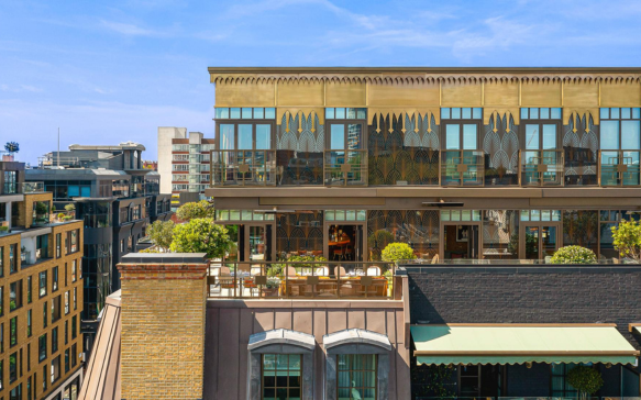 View of a rooftop restaurant or lounge with outdoor seating, string lights, and glass windows, overlooking city buildings under a blue sky.