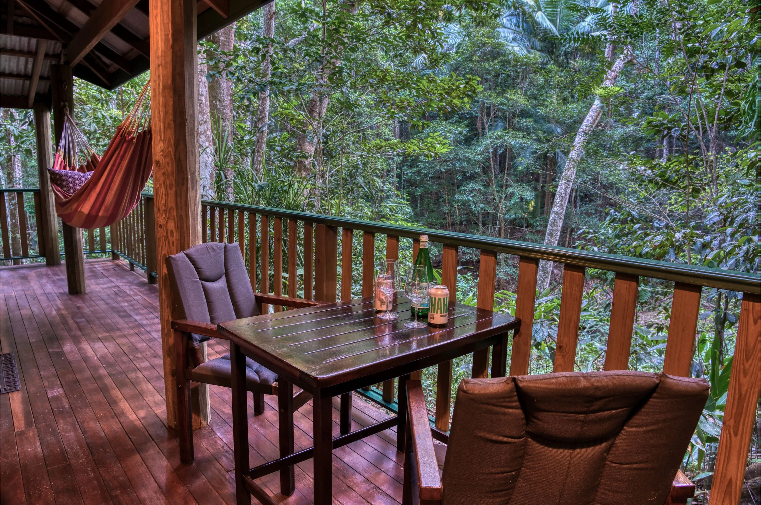 A wooden balcony with a table and chairs overlooking a lush green forest. There's a hammock hanging from the balcony's wooden beams, and a bottle of drink and glasses on the table.