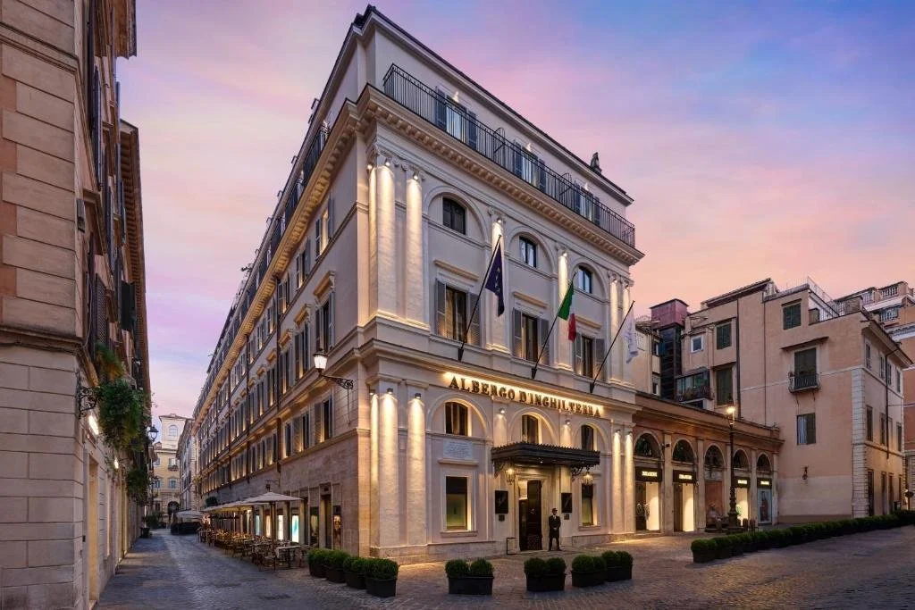 Exterior view of the Alberghetto Delle Camelie hotel at dusk in an Italian city, with flags on the facade and outdoor seating on the sidewalk.