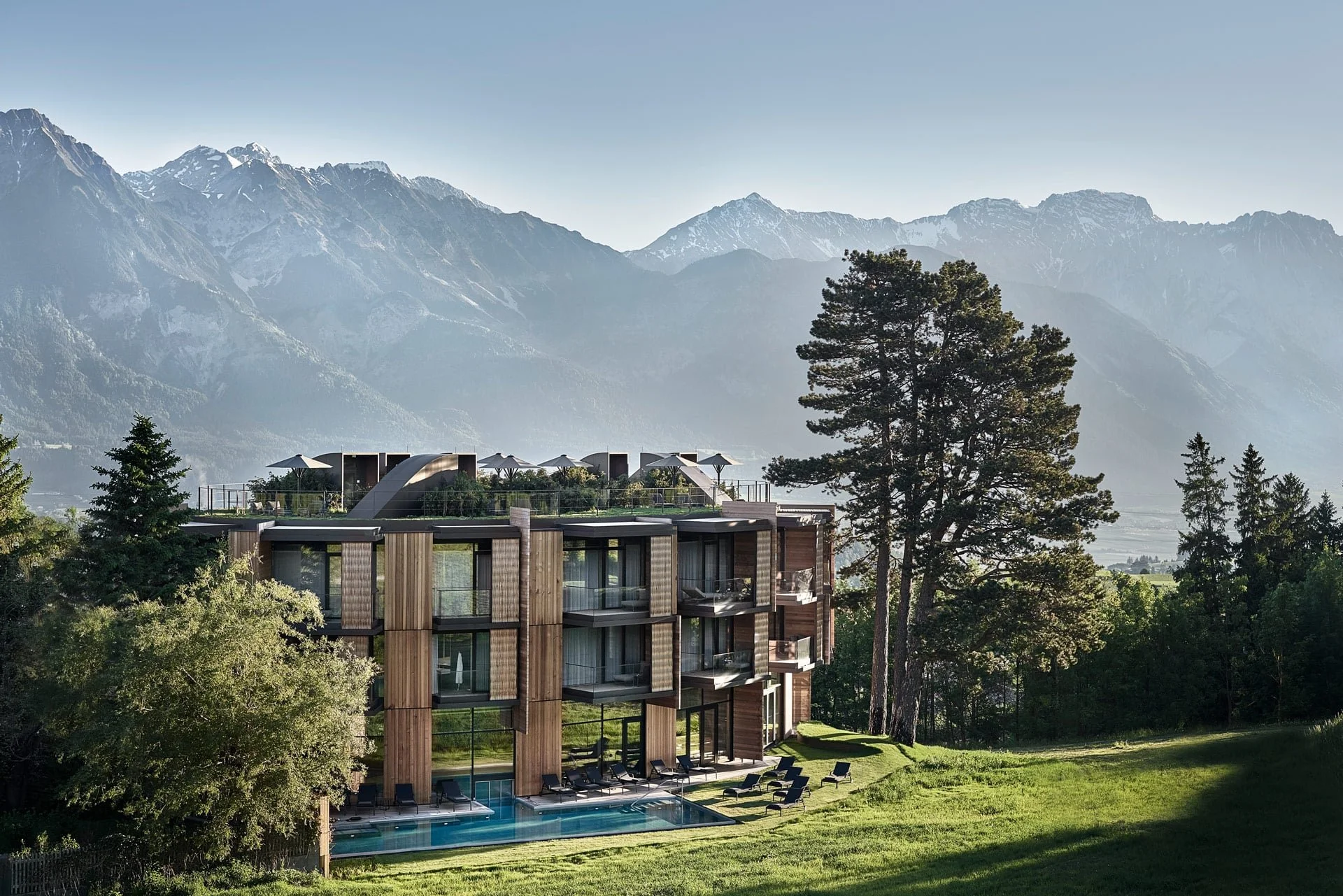Luxury modern hotel with outdoor pool, surrounded by trees, with mountains in the background.