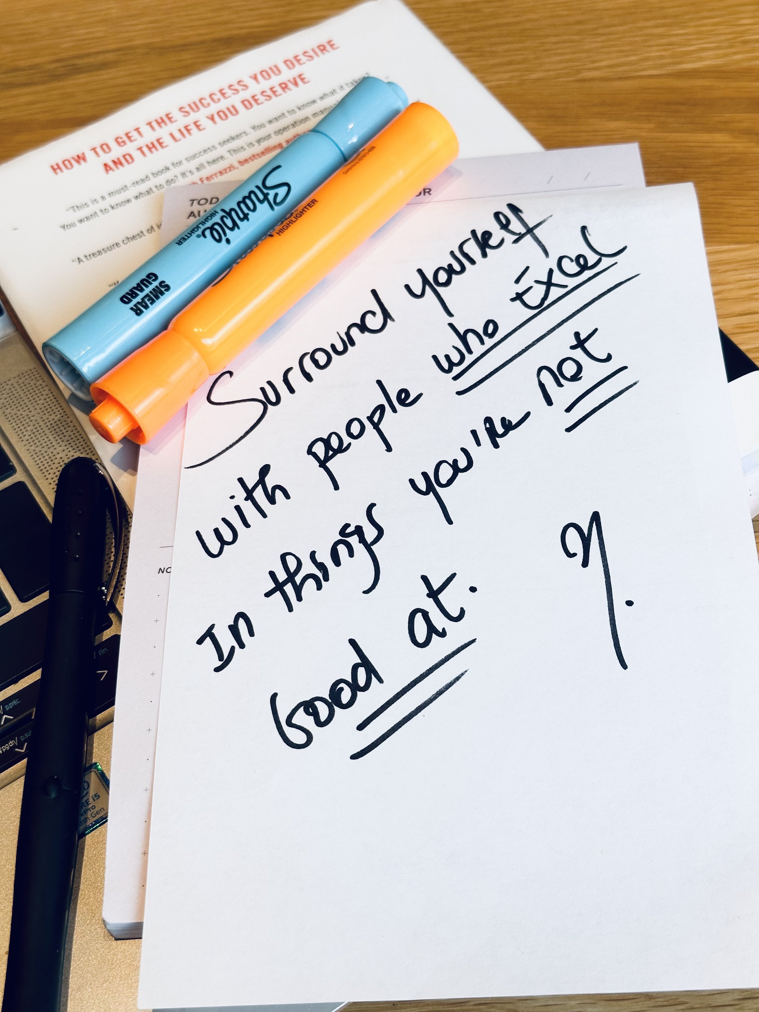 A handwritten note on a piece of paper says, 'Surround yourself with people who excel in things you're not good at.' The note is on a book with a partially visible title, accompanied by an orange and a blue Sharpie highlighter, and a black pen.