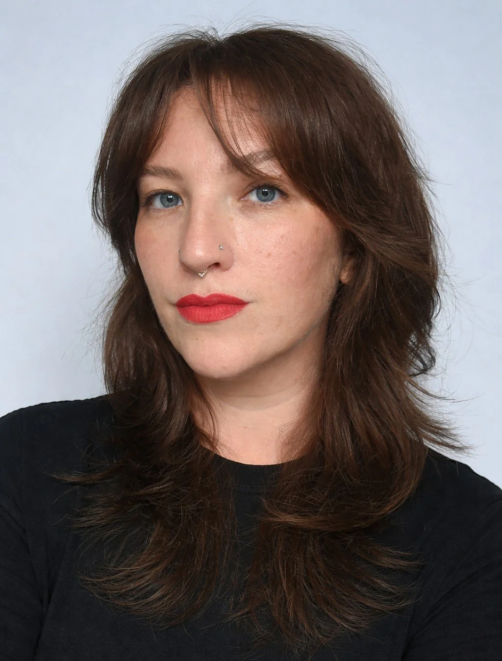 A woman with red hair, blue eyes, and fair skin, wearing red lipstick and a black top, looking at the camera against a plain light background.