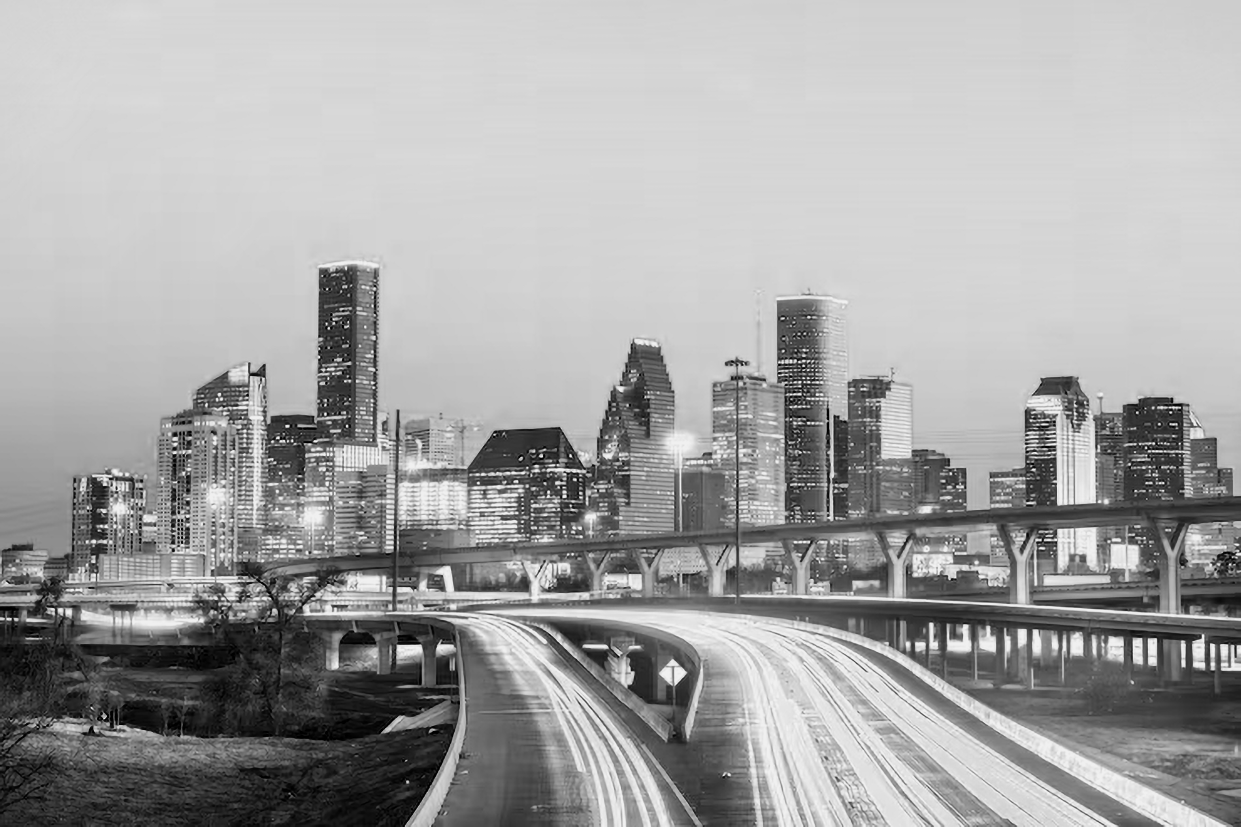 Black and white photo of a city skyline with tall buildings, a highway with streaks of light from moving vehicles, and bridges in the foreground.
