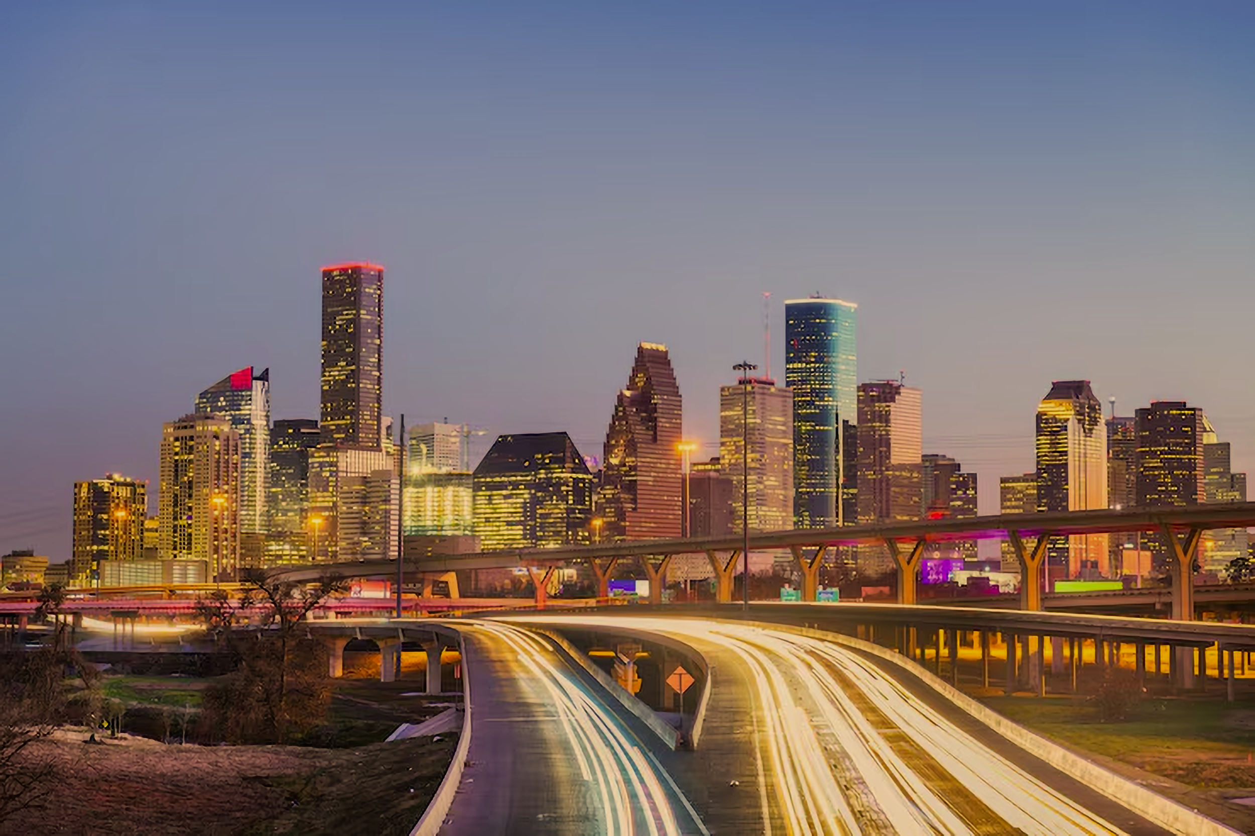 City skyline during dusk with illuminated skyscrapers, highways with light trails from moving vehicles, and a clear sky.