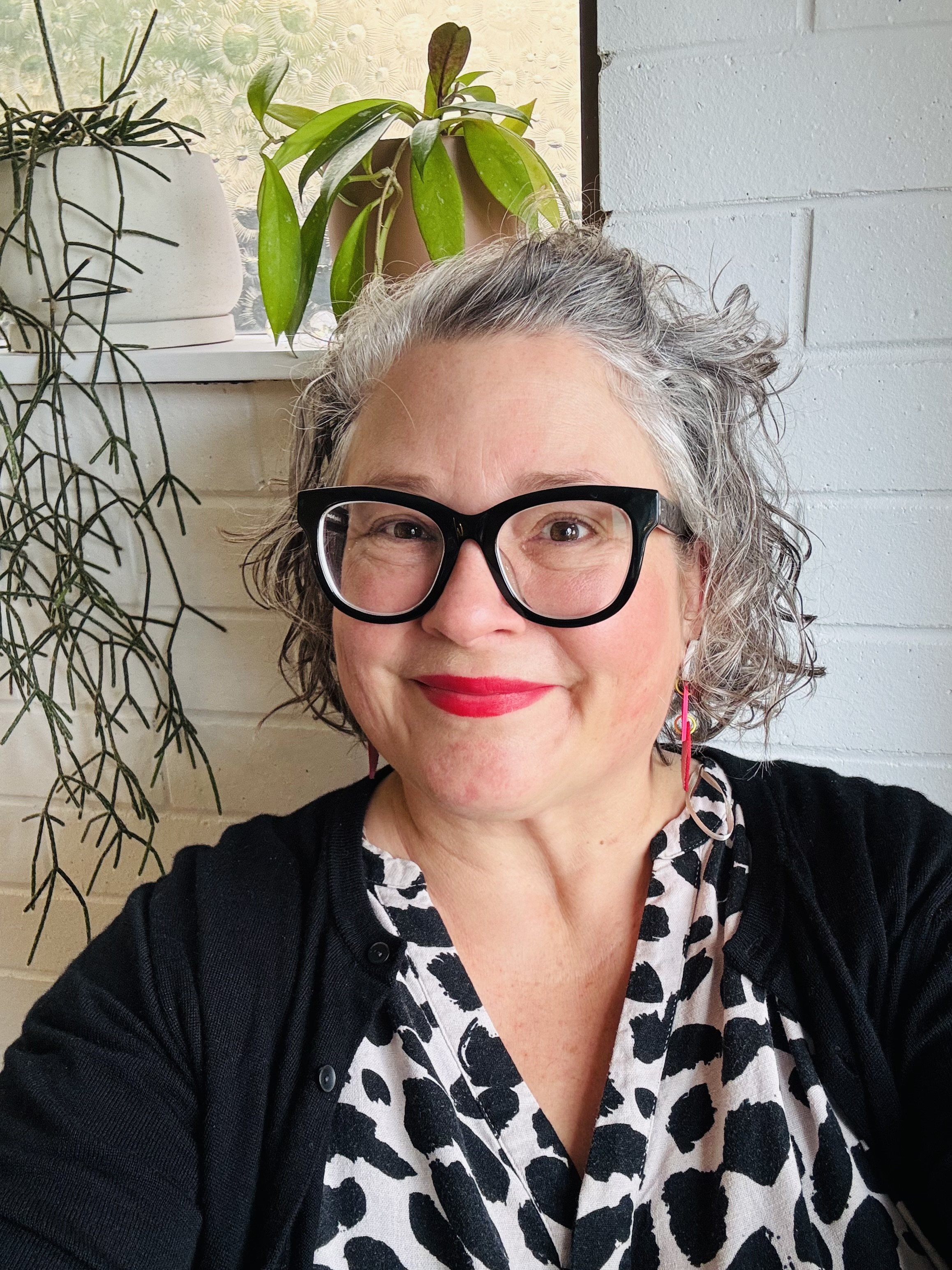 A woman with short curly gray hair, wearing black thick-framed glasses, red lipstick, a black and white animal-print dress, and a black cardigan, smiling in front of a white brick wall with indoor plants on a windowsill.