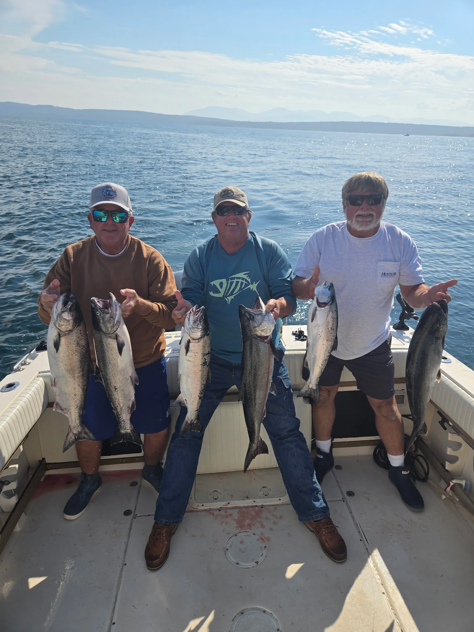 Three men on a boat holding large salmon they caught, with water and distant land in the background, on a sunny day.