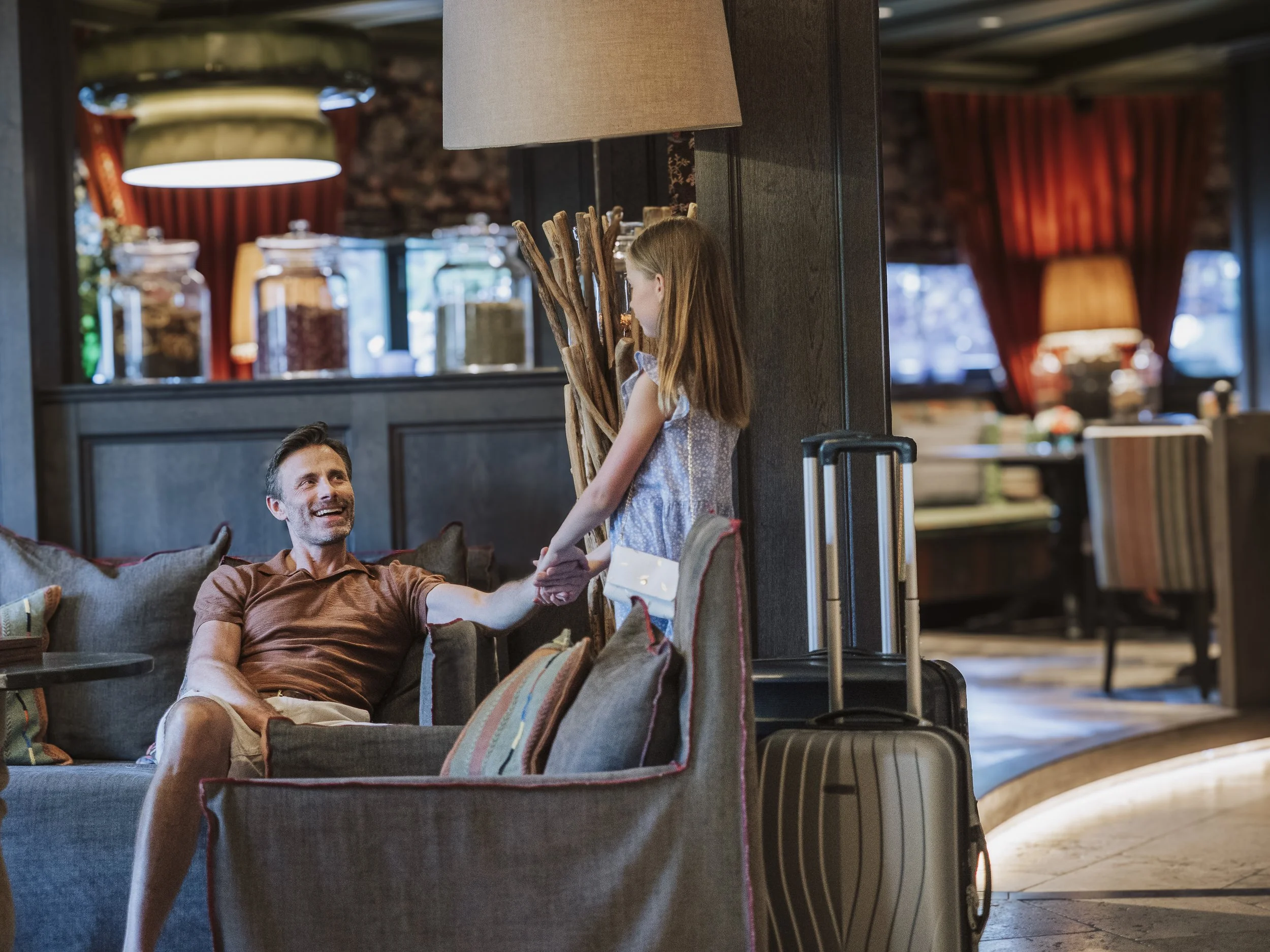 Ein Mann im Wohnzimmer schüttelt einem Mädchen die Hand, während sie ihre Handlungen umfassen. Das Mädchen hat einen Koffer neben sich, und im Hintergrund gibt es einen Raum mit großen Glasgefäßen und warmem Licht.