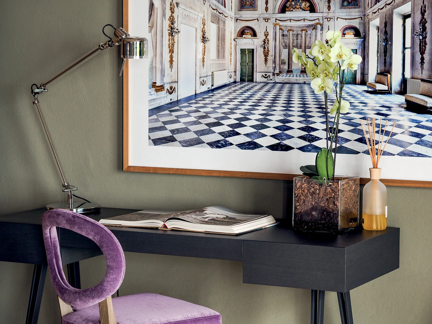 Ein schwarzer Schreibtisch mit einer offenen Zeitschrift, einer Tischlampe, einem Glasvasen mit einer Orchidee und einem Fläschchen mit Diffuser, vor einer Wand mit einem großen Wandbild eines eleganten Ballsaal mit schwarzem und weißem Fliesenboden 