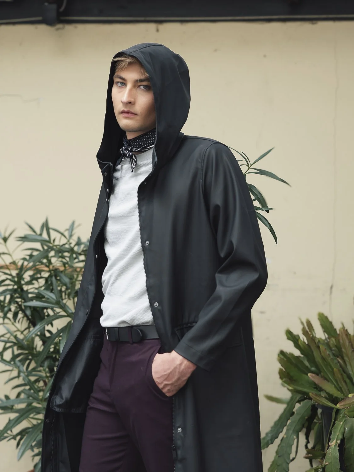 Ein junger Mann trägt einen schwarzen Kapuzenmantel, eine weiße Hemd und dunkle Hosen, steht vor einer Wand mit Pflanzen im Hintergrund.