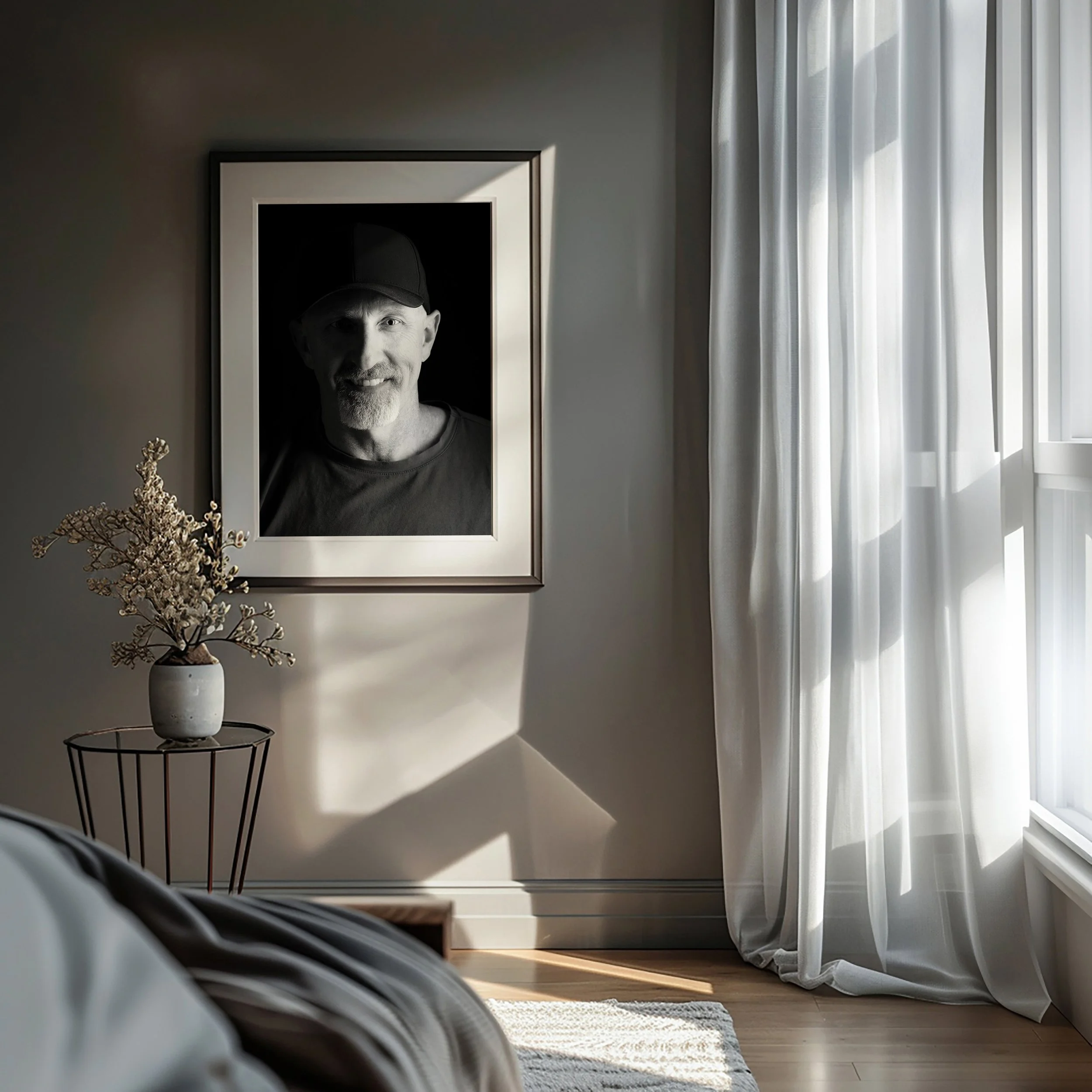 Schwarze und weiße Fotografie eines Mannes in einem Bilderrahmen an einer Wand, neben einem Fenster mit weißen Vorhängen, auf einem kleinen Tisch eine Vase mit Trockenblumen.