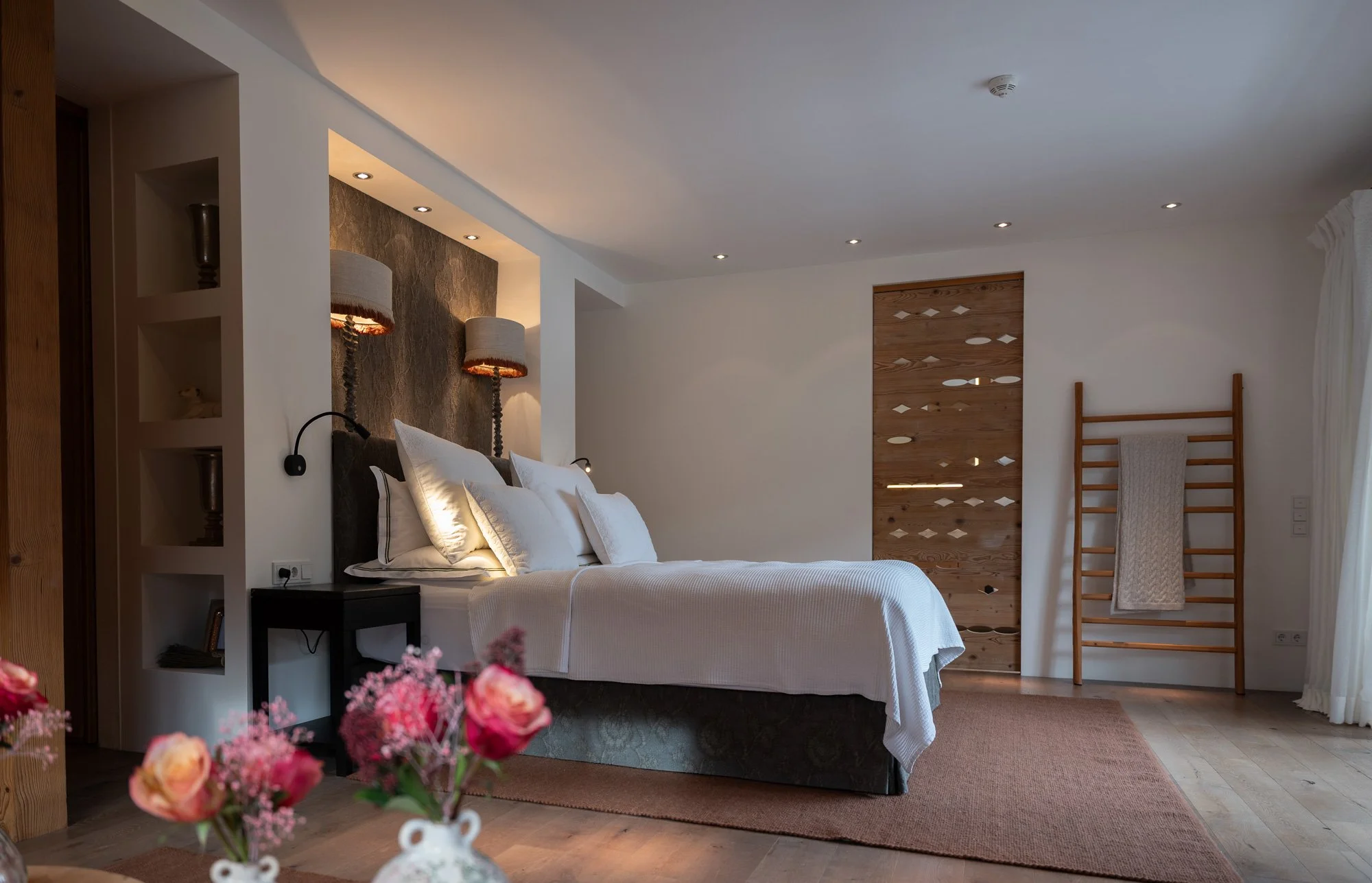 Ein modern gestaltetes Schlafzimmer mit weißem Bett, Wandlampen, Holzdekor, einem Holzrollladen und einer Kleiderstange in Holzoptik.