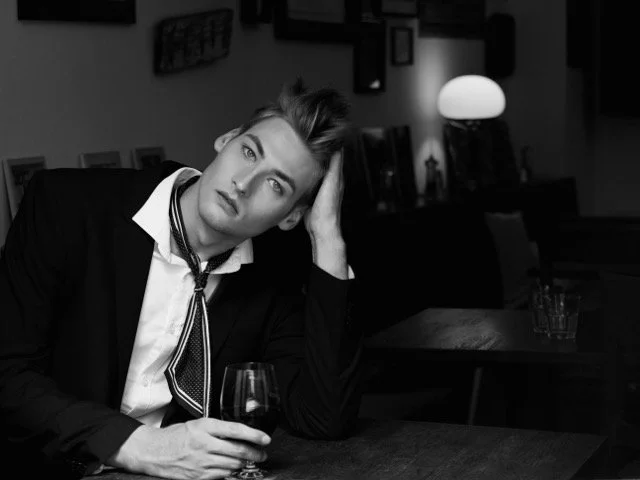 Ein junger Mann sitzt in einem Restaurant, hält ein Glas Wein in der Hand und sieht nachdenklich in die Kamera, schwarz-weiße Aufnahme.