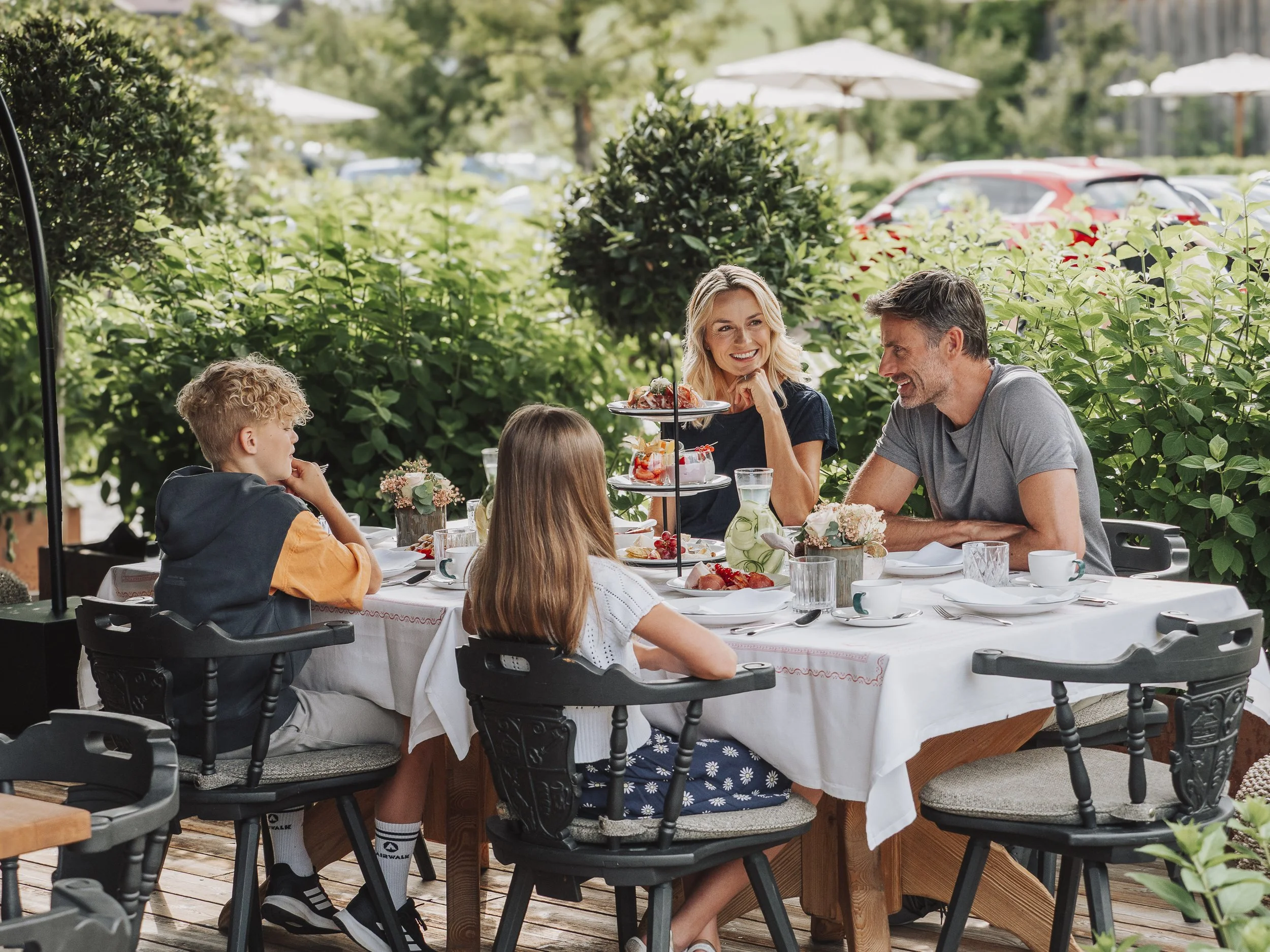 Familie beim Frühstück im Freien auf einer Terrasse, umgeben von Grün, mit Kuchen, Blumen und Geschirr auf dem Tisch.