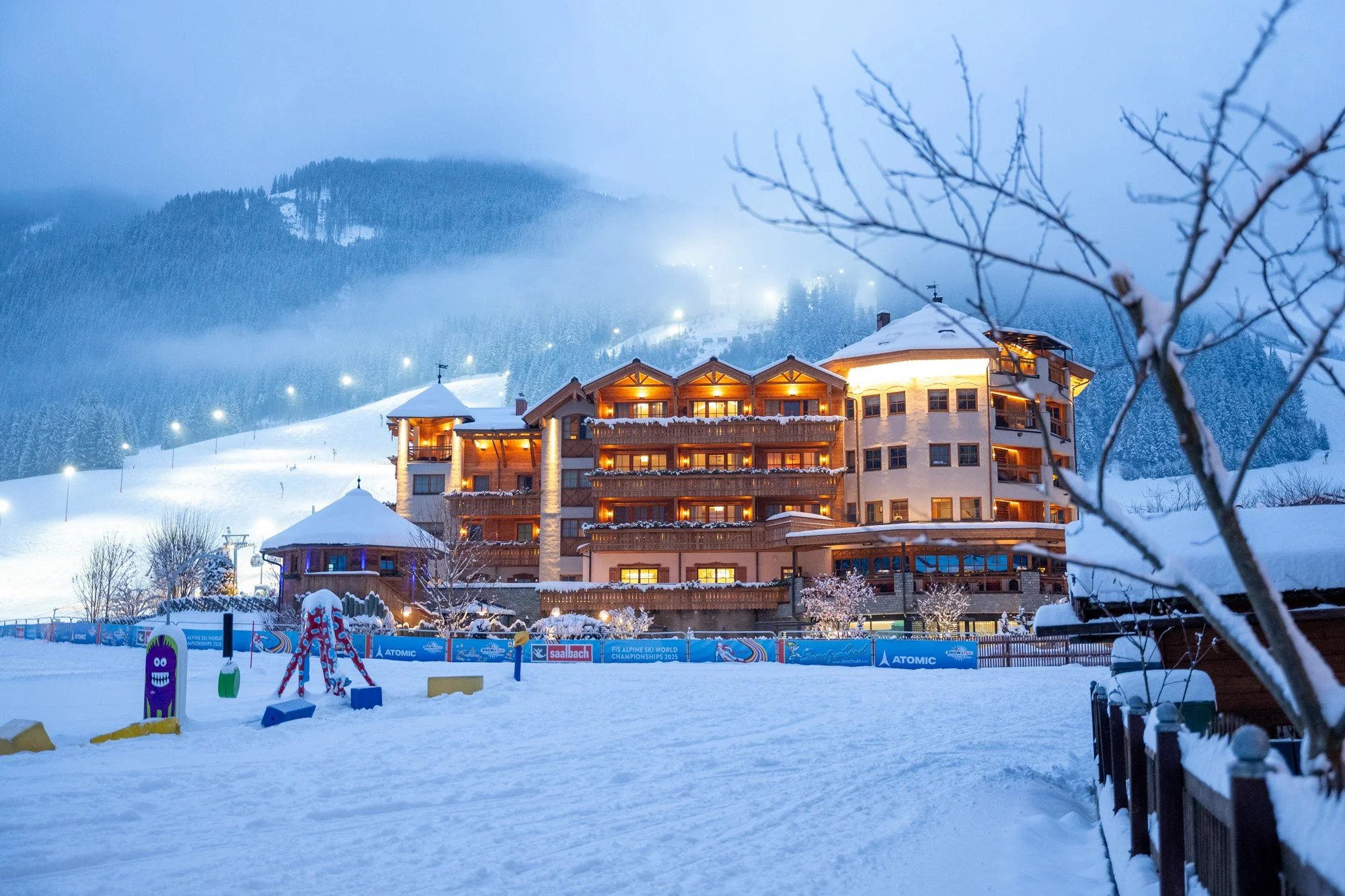 Ein schneebedeutes Hotel in den Bergen bei Nacht, beleuchtet mit warmem Licht, mit spielender Schneeschaukel im Vordergrund und bewaldeten Hügeln im Nebel im Hintergrund.