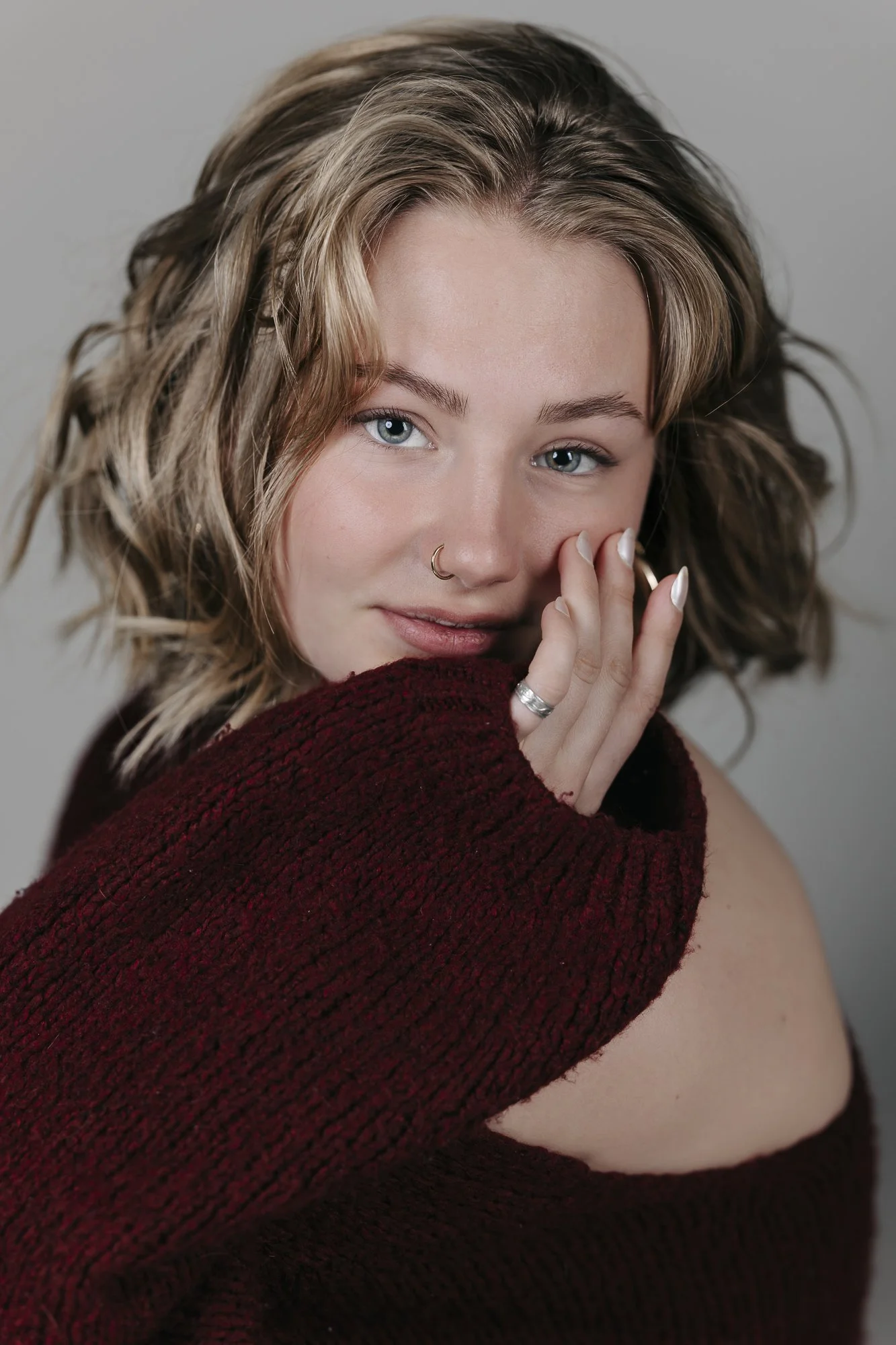 Nahaufnahme eines lächelnden jungen Frau mit blonden, lockigen Haaren, die eine Nase mit einem Ringlein und mehrere Ringe am Finger trägt. Sie trägt einen roten Pullover mit weitem Ansatz.