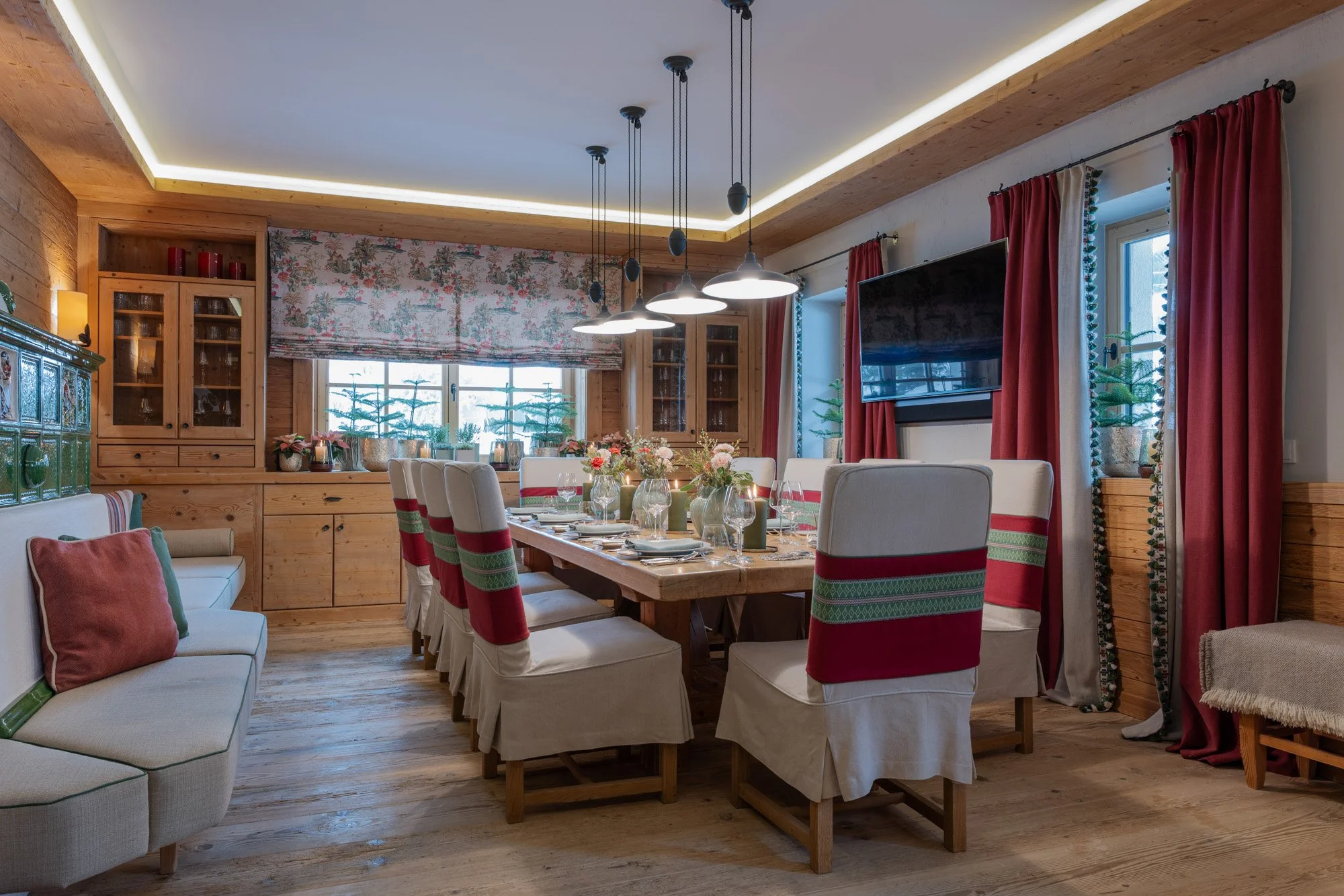 Ein festlich gedeckter Essentisch in einem rustikalen Holzzimmer mit schnurgebundenen roten, weißen und grünen Stuhlhussen, Blumen, Kerzen und großen Fenstern mit roten Vorhängen.