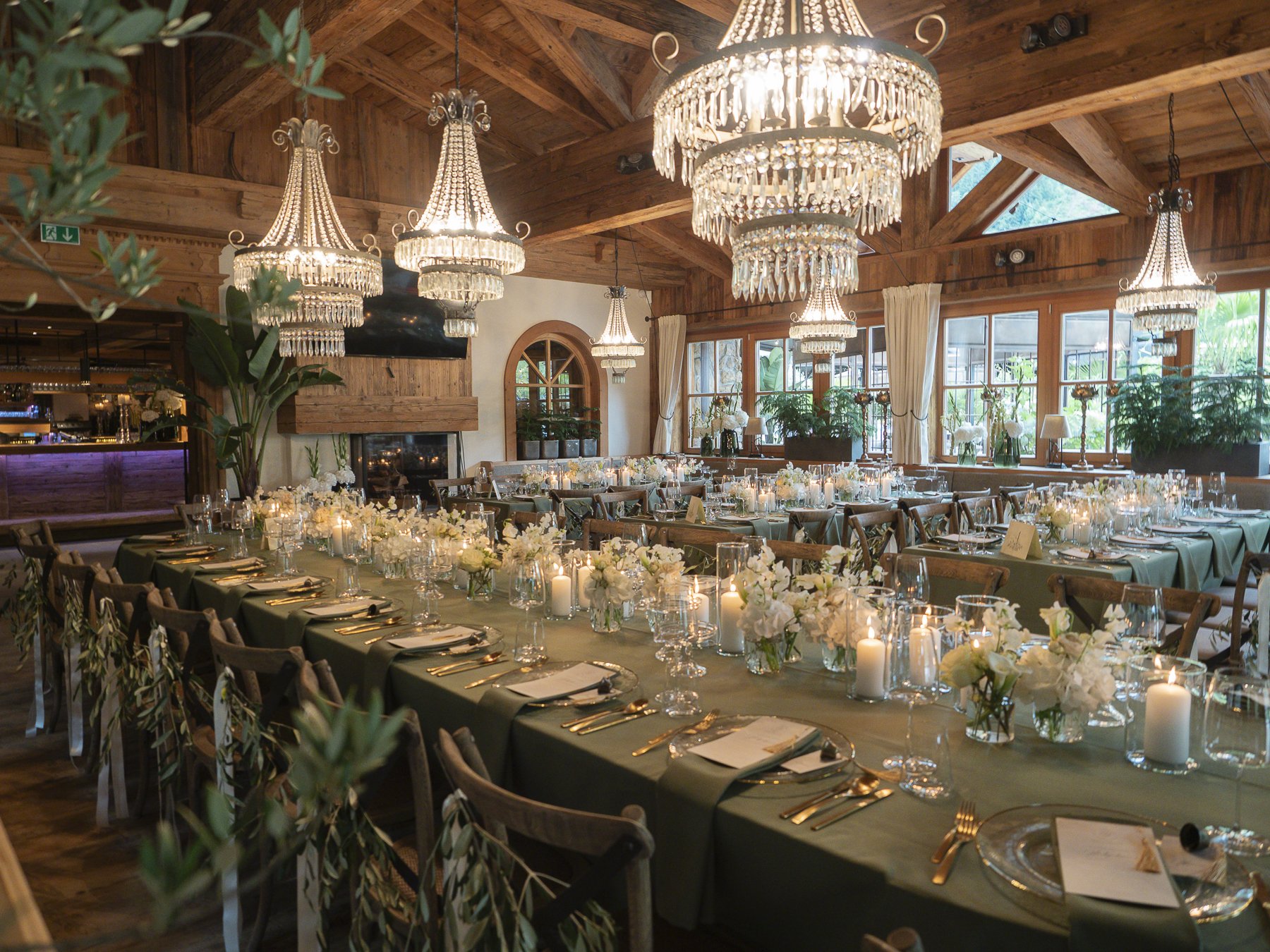 Ein festlich gedeckter Saal mit langen Tischen, geschmückt mit weißen Blumen, Kerzen und goldenen Bestecken. Deckenlampen und viel Tageslicht durch große Fenster. Holzdecke und -wände, viel Grünpflanzen im Raum.
