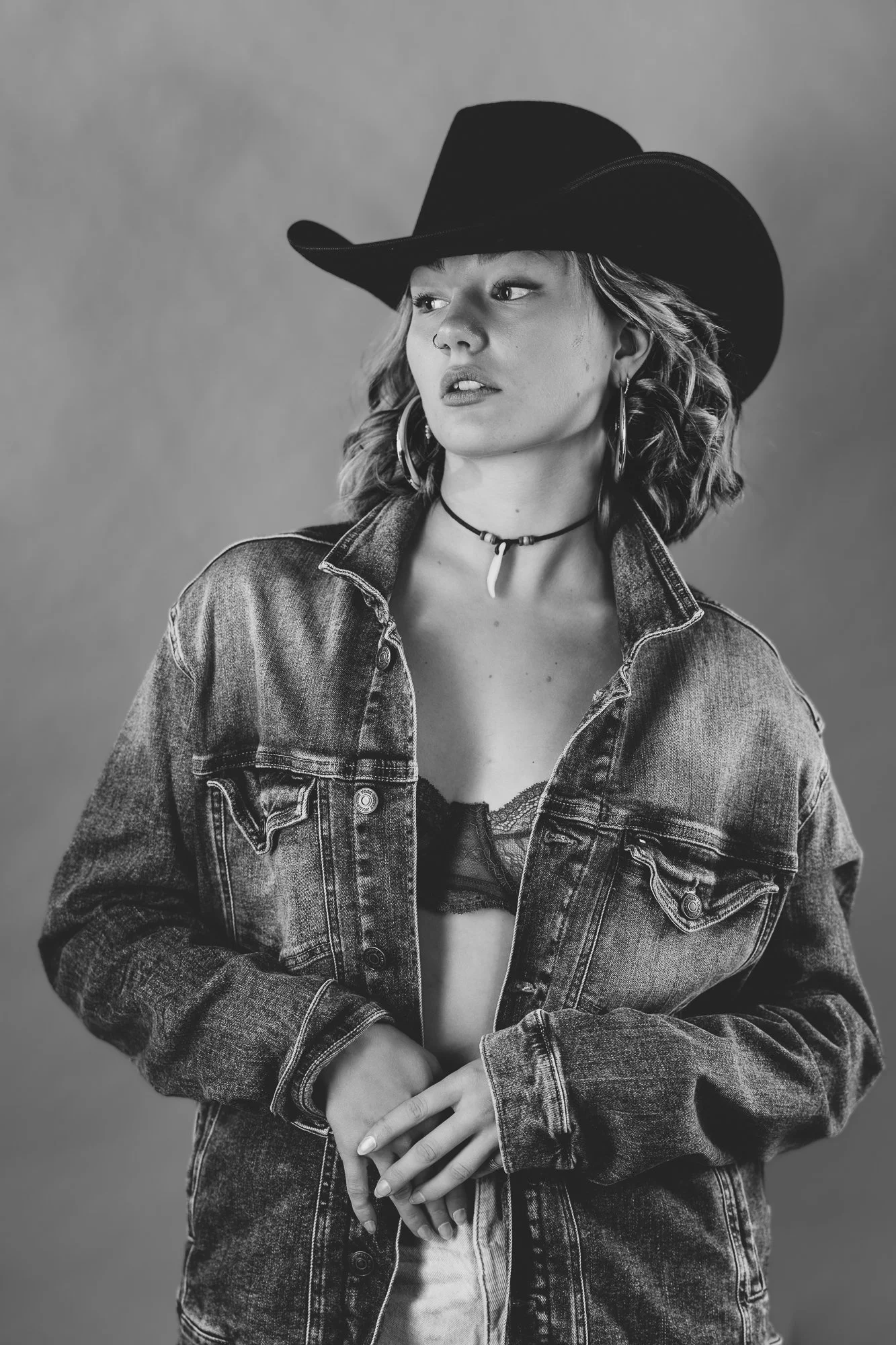 Schwarz-weiß-Foto einer jungen Frau mit Cowboyhut, Jeansjacke, Lederkorset, Halsband mit Zahnanhänger, großen Ohrringen und kurzen, lockigen Haaren.