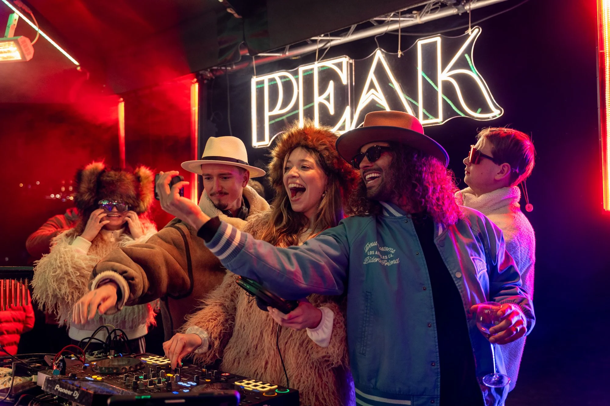 Eine Gruppe von Menschen, die auf einer Party tanzen und Spaß haben, unter Neonlichtern, mit einigen DJ-Equipment im Vordergrund.