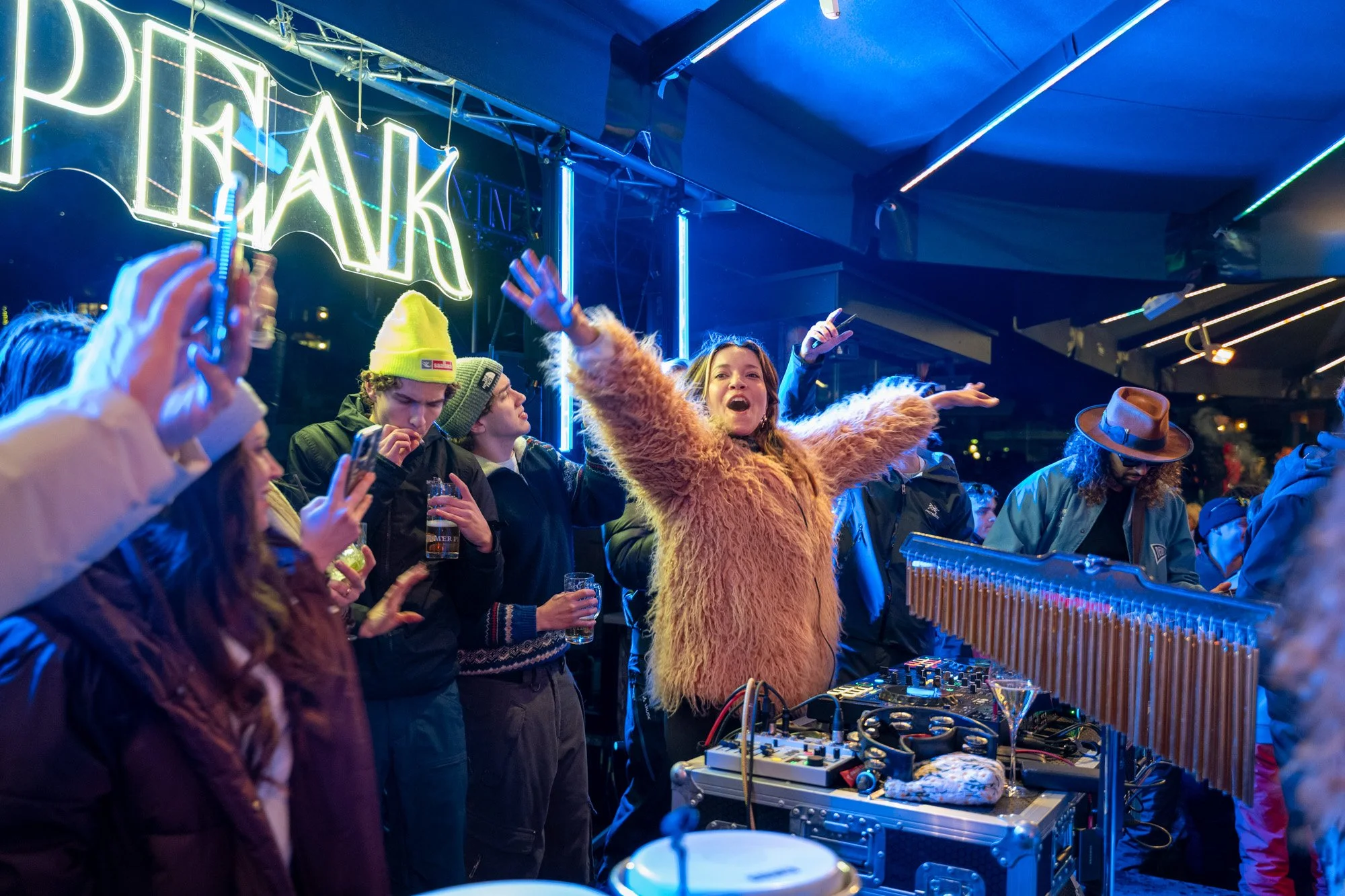 Menschen feiern bei einer Party oder einem Konzert, mit einem DJ, der auflegte. Die Szene ist bunt beleuchtet, mit einem Neon-Schild im Hintergrund, das auf Englisch "PEAK" steht.
