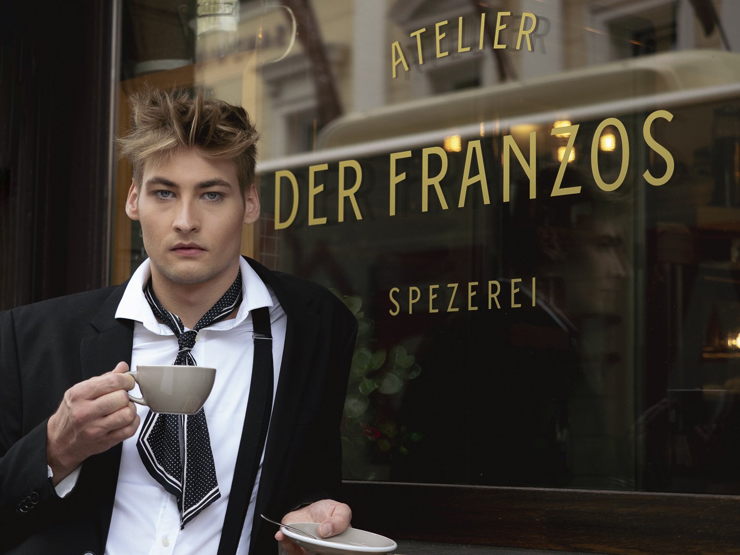 Ein junger Mann mit zerzaustem Haar, weiße Hemd, schwarze Jacke, und einer schwar-weißen Krawatte, trinkt aus einer Tasse, vor einem Schaufenster eines italienischen Restaurants mit der Aufschrift "Atelier der Französischen Spezereien" auf Deutsch.