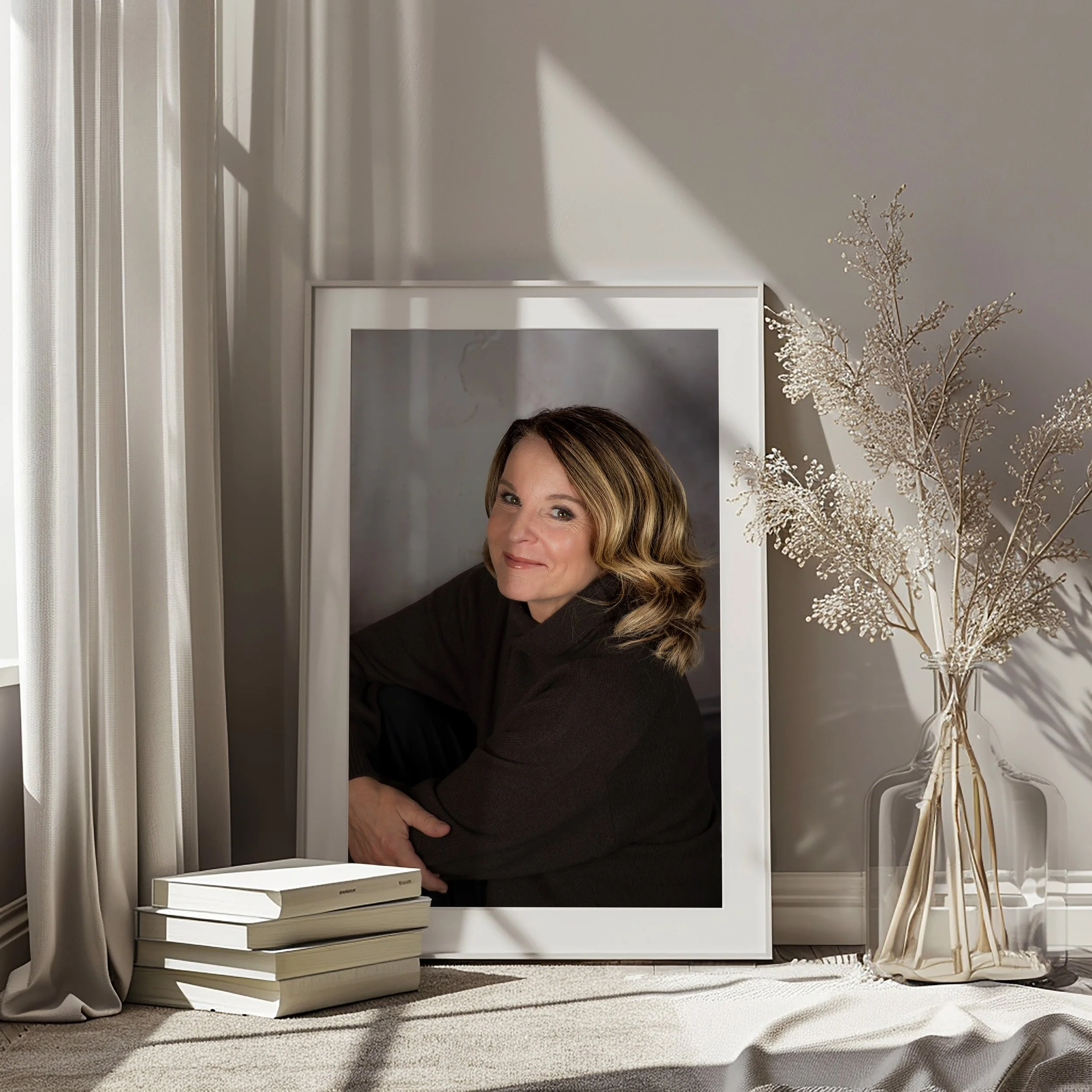 Ein Portrait einer Frau, die in einem weißen Rahmen Fotos fotografiert ist, in einem hellen Raum mit Sonnenlicht und einem weißen Vorhang, neben einer Vase mit Trockenblumen und einem Stapel Bücher.