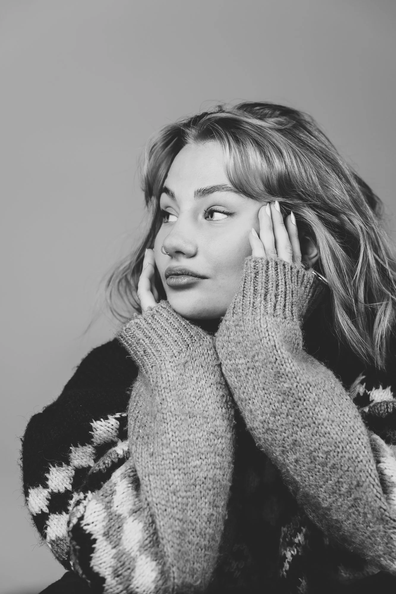 Schwarz-weiße Porträtaufnahme einer jungen Frau mit Schulterlänge Haaren, die eine graue, dicke, gestrickte Pullover trägt, die ihre Hände auf das Gesicht stützt, blickt seitlich, neutraler Gesichtsausdruck.