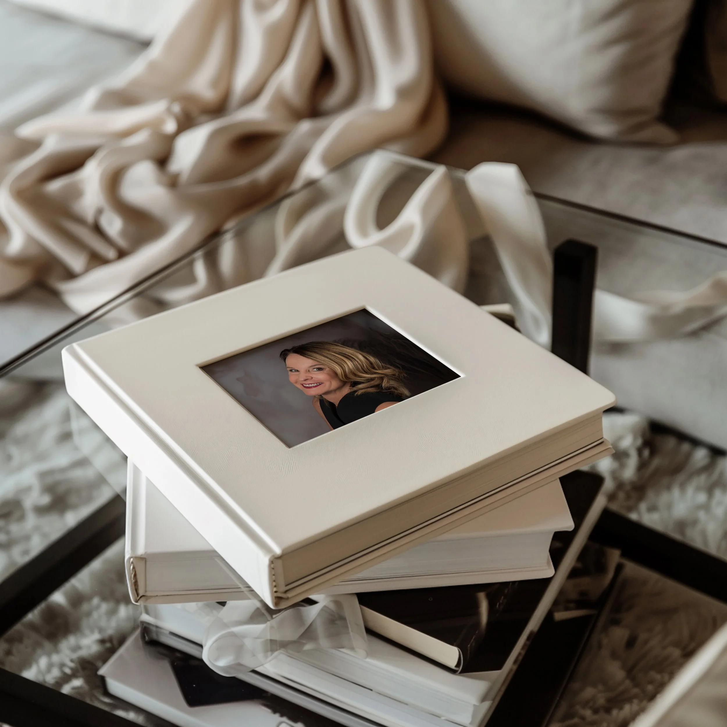 Ein aufgeschlagenes Fotobuch mit einem Porträt einer lachenden Frau liegt auf einem Stapel anderer Bücher auf einem Couchtisch, im Hintergrund ist ein weiches Kissen und Stoff. Die Bildqualität ist hochauflösend, mit natürlichen Farben.